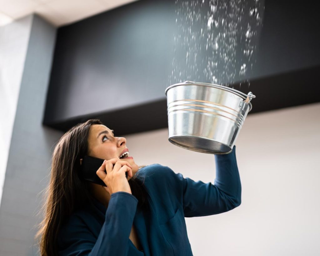 Worried woman on cell phone holding silver bucket underneath leaking ceiling